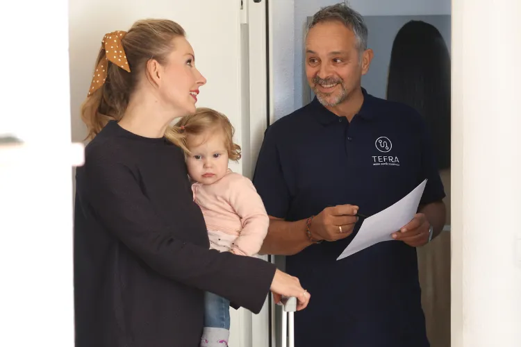 Zwei Personen an einer Tür, die eine ist eine Frau in schwarzem Shirt und hält ein Kind auf dem Arm. Die andere Person ist ein Mann mit einem Blatt Papier in der Hand und einem dunkelblauen T-Shirt mit TEFRA Logo.