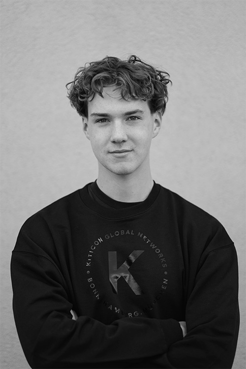 Porträtfoto von Julian Kenfenheuer, Teammitglied bei KITICON Global Networks, mit lockigem Haar und schwarzem Pullover, der das KITICON-Logo trägt.